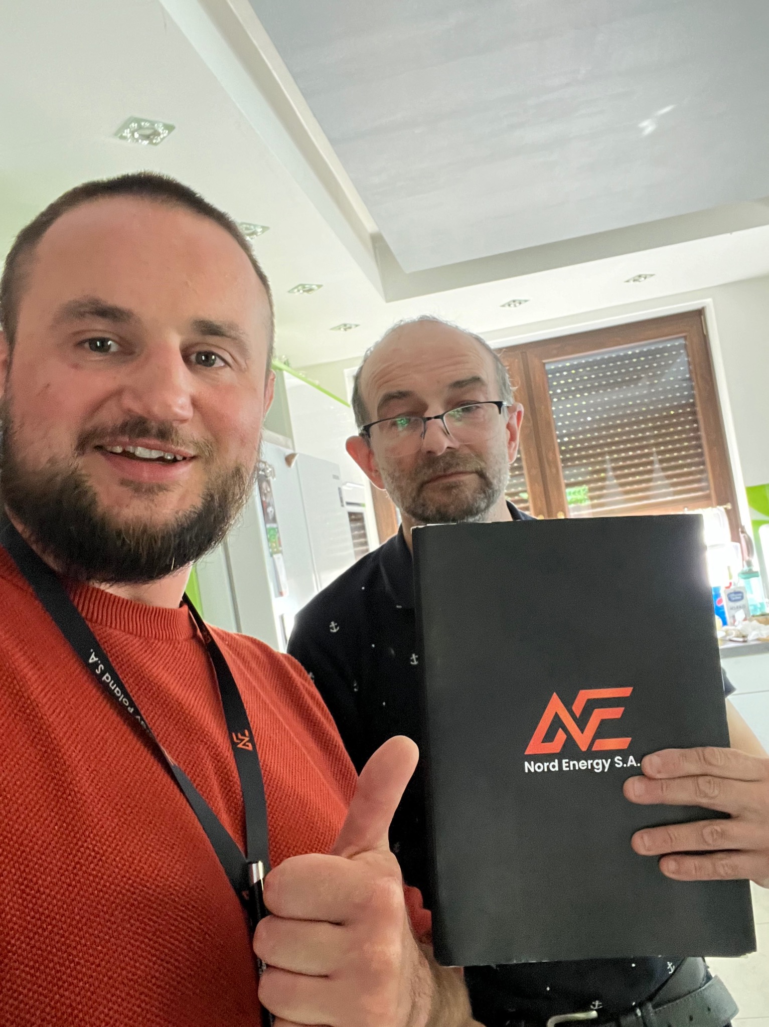 Dwóch mężczyzn, jeden z brodą w pomarańczowym swetrze z identyfikatorem Poland S.A., drugi z okularami trzyma czarny folder z logo Nord Energy S.A. i daje kciuk w górę, w tle kuchnia.