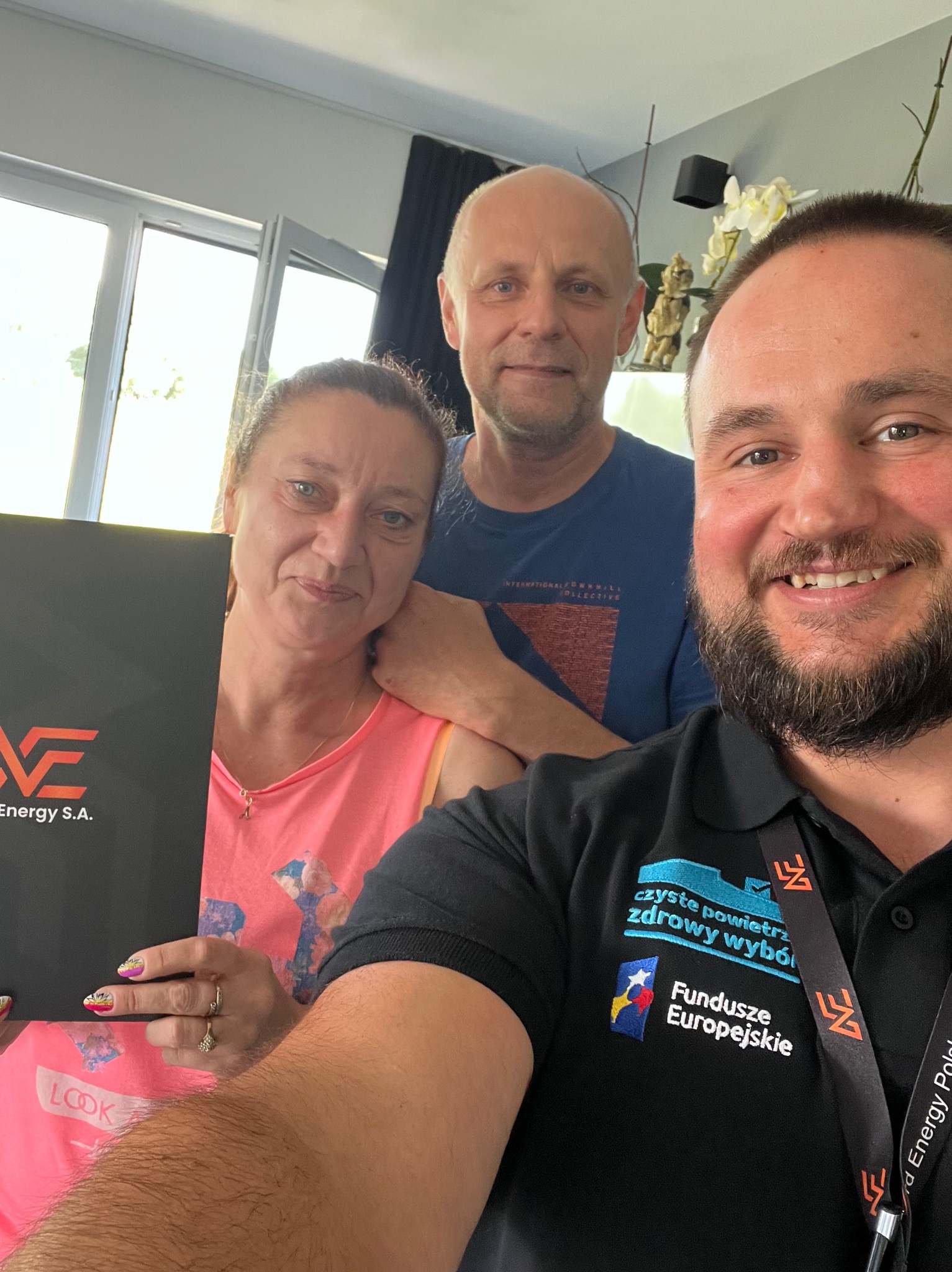 Trzy osoby pozują do zdjęcia w pomieszczeniu, kobieta trzyma czarną teczkę z logo firmy Energy S.A., pracownik firmy w czarnej koszulce z logo Funduszy Europejskich robi selfie.
