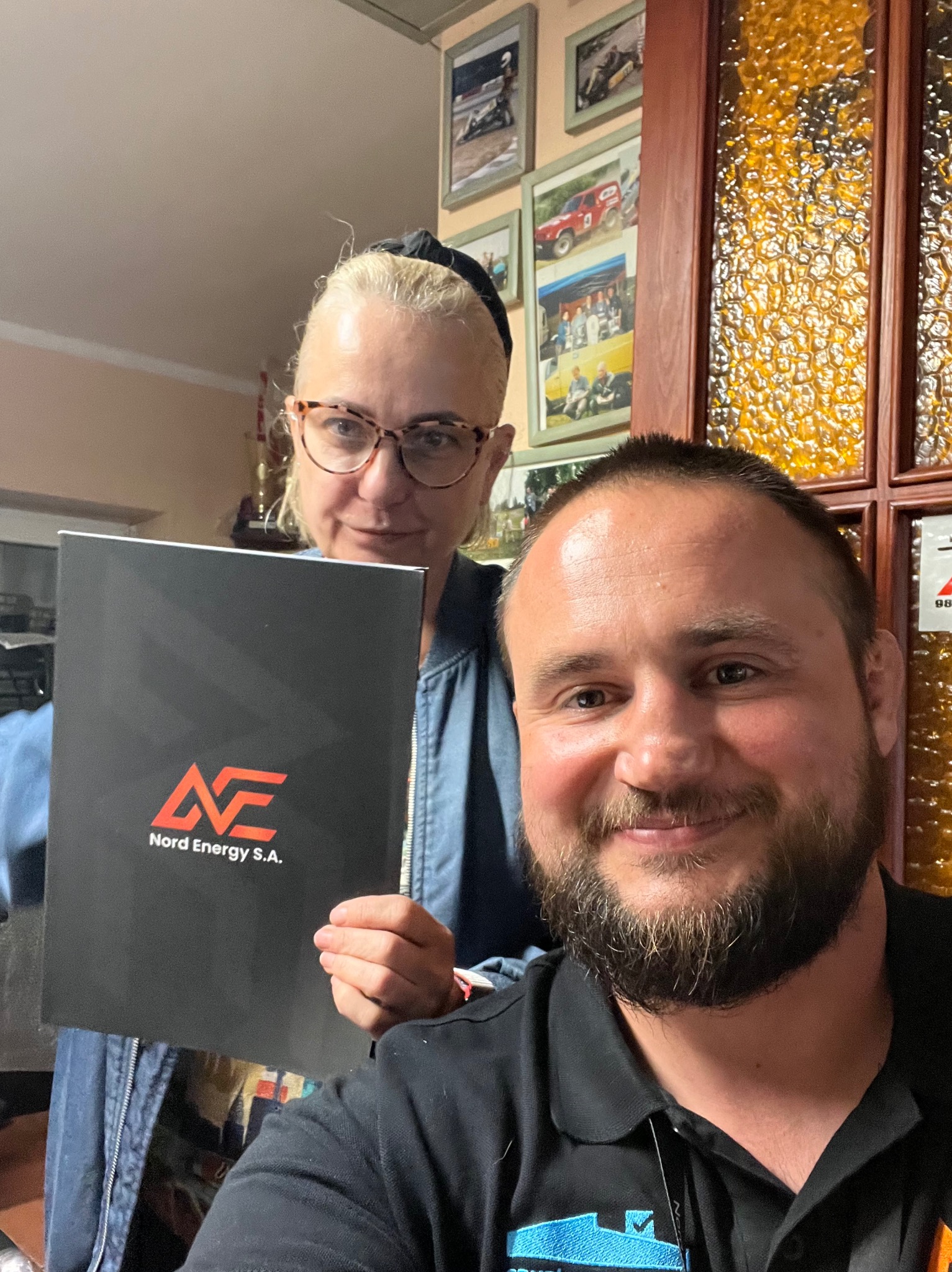 Dwie osoby prezentują czarny folder z logo Nord Energy S.A. na tle ściany z ramkami zdjęć i dekoracyjną szybą.