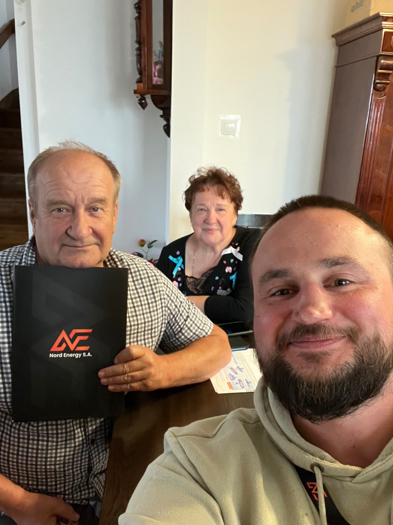Trzy osoby siedzą przy stole, mężczyzna z przodu robi selfie, starszy mężczyzna trzyma czarny folder z logo Nord Energy S.A., w tle kobieta.