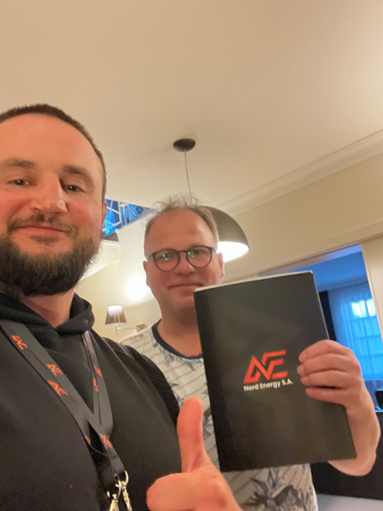 Selfie dwóch mężczyzn z folderem Nord Energy S.A. w dłoni jednego z nich, widoczny kciuk w górę i smycz z logo firmy.
