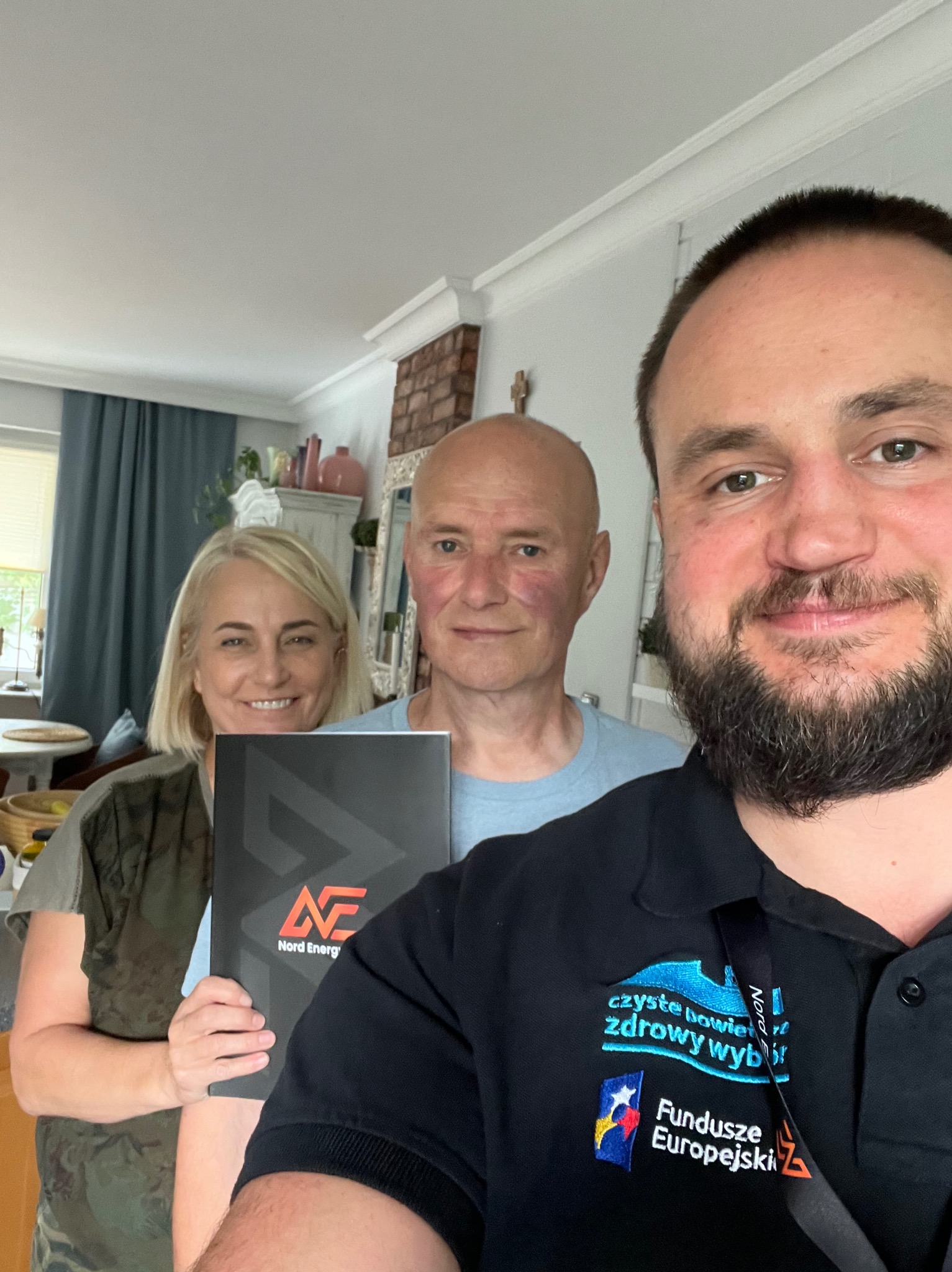 Trzy osoby, w tym pracownik firmy Nord Energy, prezentują folder z logo firmy w jasnym wnętrzu domu.
