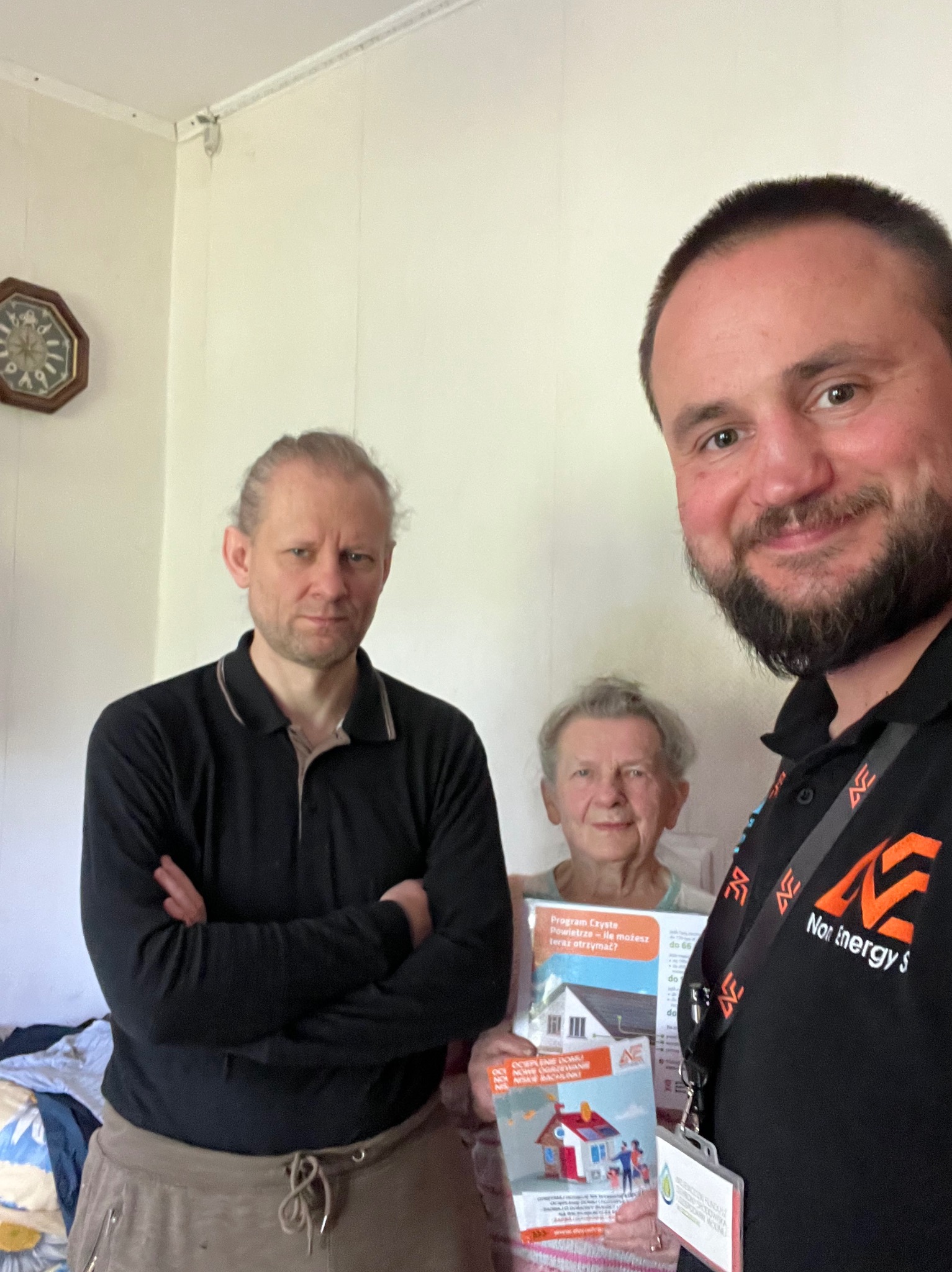 Zdjęcie zrobione z bliska przedstawia pracownika firmy Nor Energy S. z ulotkami programu 'Czyste Powietrze', pozującego z dwoma osobami wewnątrz domu. Ulotki zawierają wizualizacje domów...