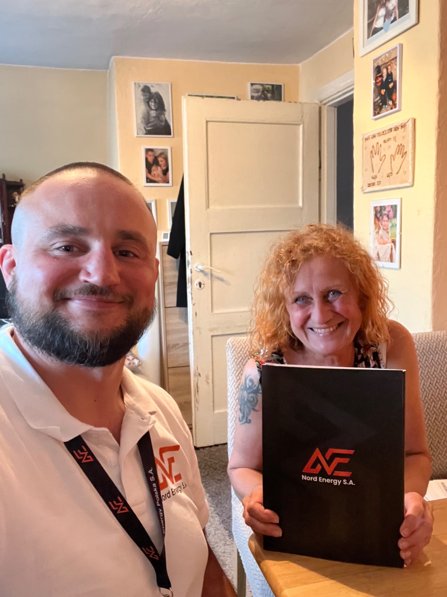 Uśmiechnięta kobieta z tatuażem na ramieniu trzyma czarne pudełko z logo Nord Energy S.A., obok mężczyzna z identyfikatorem tej samej firmy, w tle zdjęcia rodzinne na ścianie.