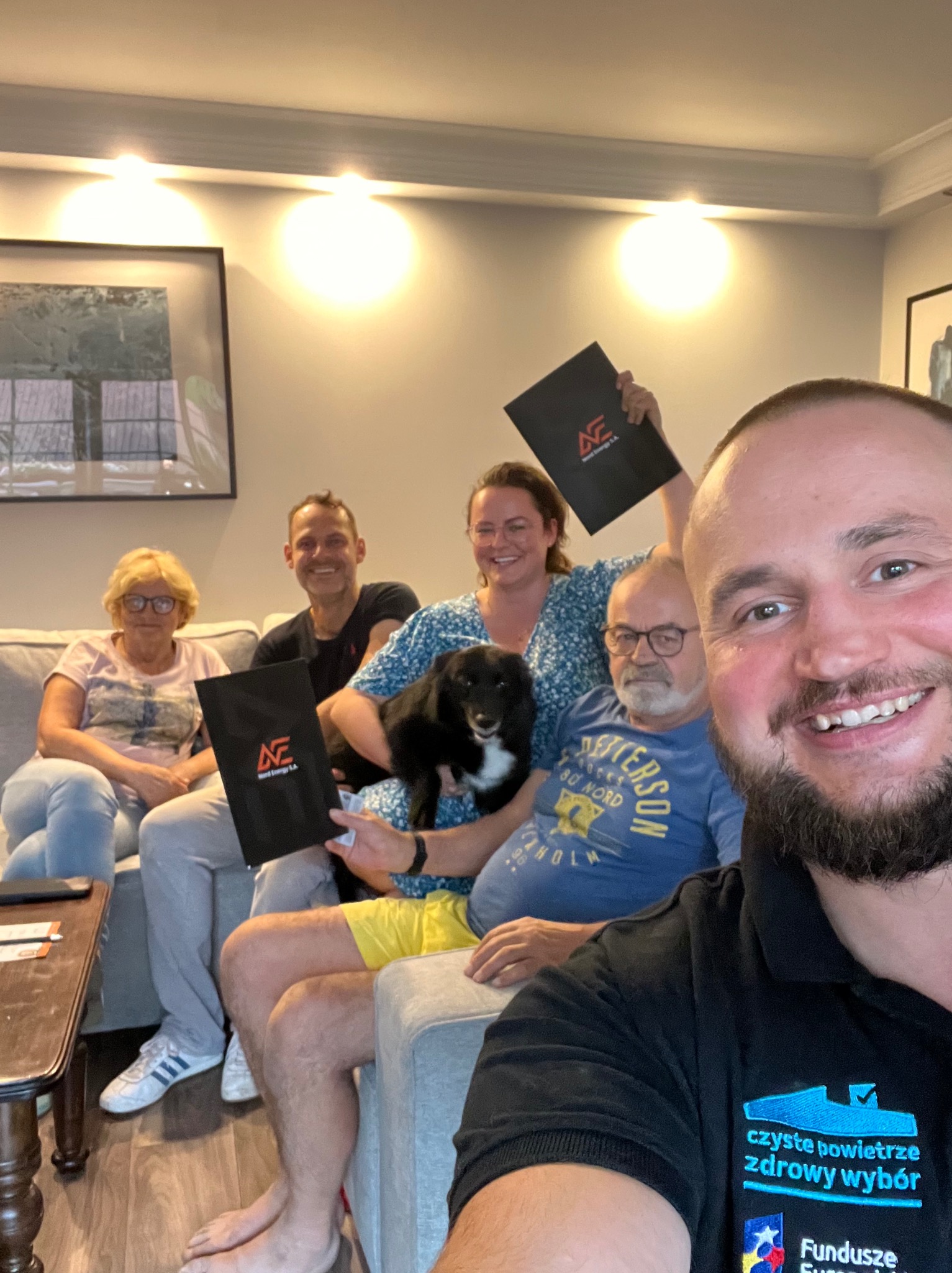 Grupa ludzi siedzi na kanapie, trzymając czarne teczki z logo firmy, z uśmiechem pozując do zdjęcia selfie z pracownikiem w koszulce z logo programu 'Czyste Powietrze'.