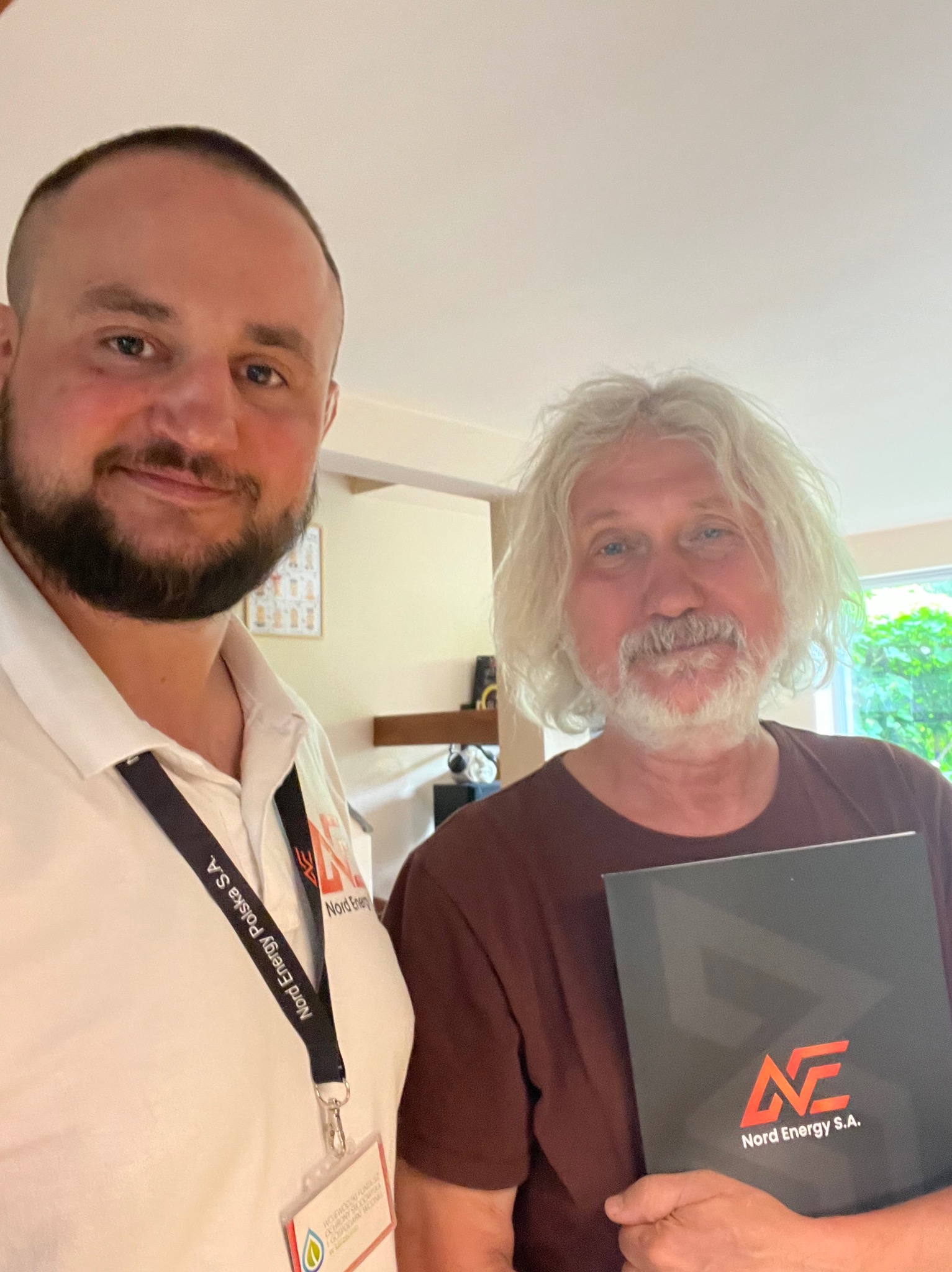 Mężczyzna w koszuli polo z identyfikatorem firmy Nord Energy Polska S.A. robi sobie selfie z klientem trzymającym folder z logo Nord Energy S.A.
