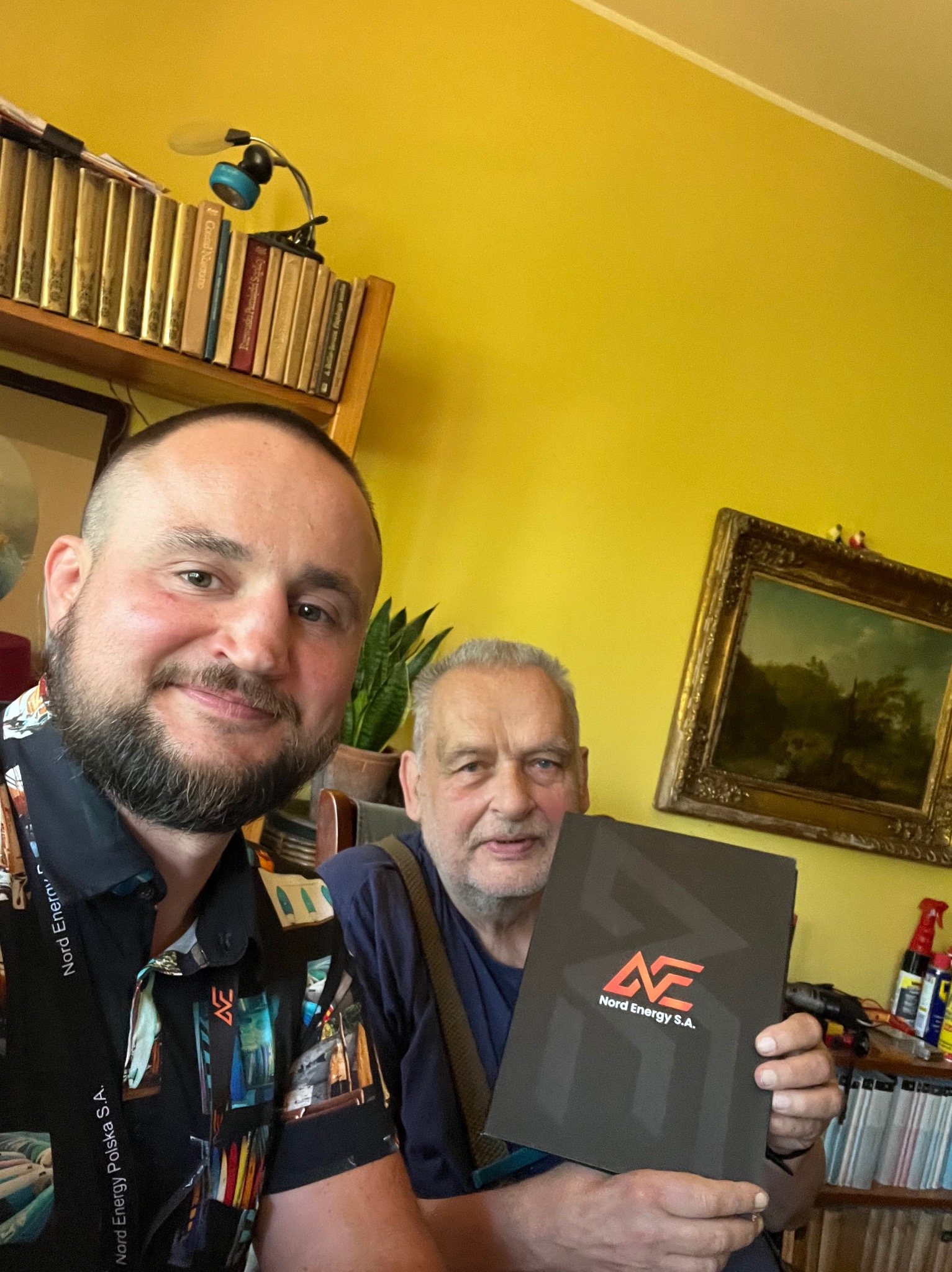 Mężczyzna w koszuli z logo Nord Energy Polska S.A. robi selfie z klientem trzymającym w dłoniach czarne pudełko z logo Nord Energy S.A. na tle żółtej ściany i regału z książkami.