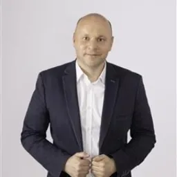 Mężczyzna w granatowej marynarce i białej koszuli, z delikatnym uśmiechem, na jasnym tle. Widoczne dłonie trzymające klapy marynarki.