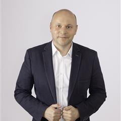 Mężczyzna w granatowej marynarce i białej koszuli, z delikatnym uśmiechem, na jasnym tle. Widoczne dłonie trzymające klapy marynarki.