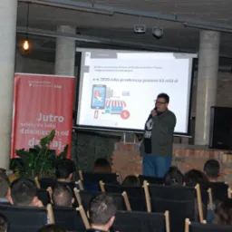 Prelegent z mikrofonem prezentuje dane o wzroście e-commerce na projektorze podczas konferencji, widownia siedzi na krzesłach.