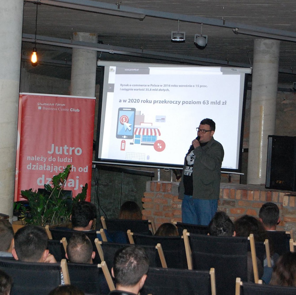 Prelegent z mikrofonem prezentuje dane o wzroście e-commerce na projektorze podczas konferencji, widownia siedzi na krzesłach.