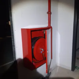 Czerwona szafka hydrantowa z rozłożonym wężem na ścianie wewnątrz budynku, z widoczną rurą doprowadzającą wodę.