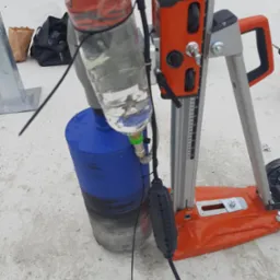 Wiertnica diamentowa z systemem chłodzenia wodą z butelki PET, pomarańczowa podstawa z regulacją wysokości, widoczna skala pomiarowa, na betonowej powierzchni.