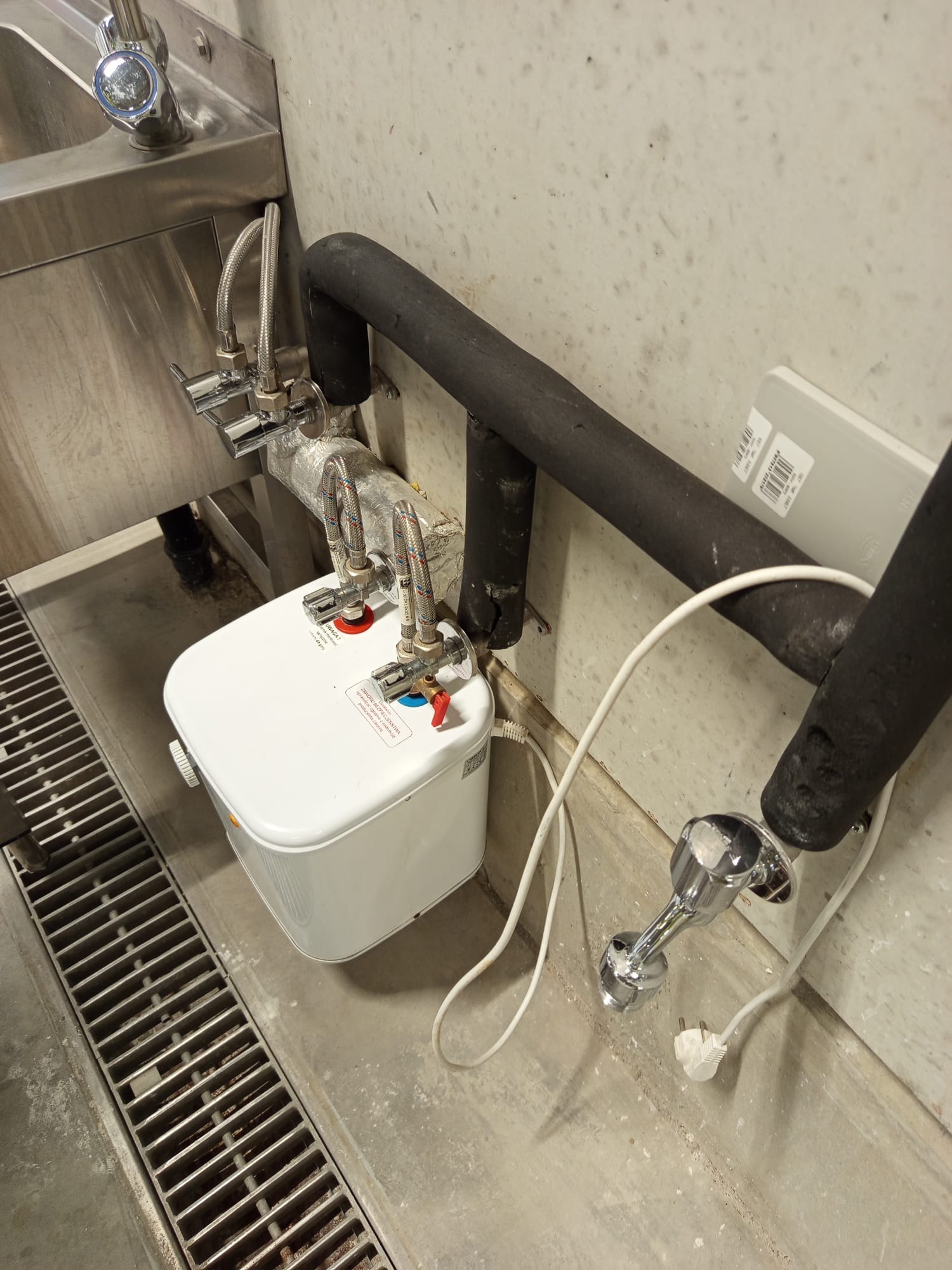 Mały, biały podgrzewacz wody zamontowany pod zlewem ze stali nierdzewnej, widoczne rury z izolacją termiczną, przyłącza wody z zaworami, oraz kabel zasilający z wtyczką.