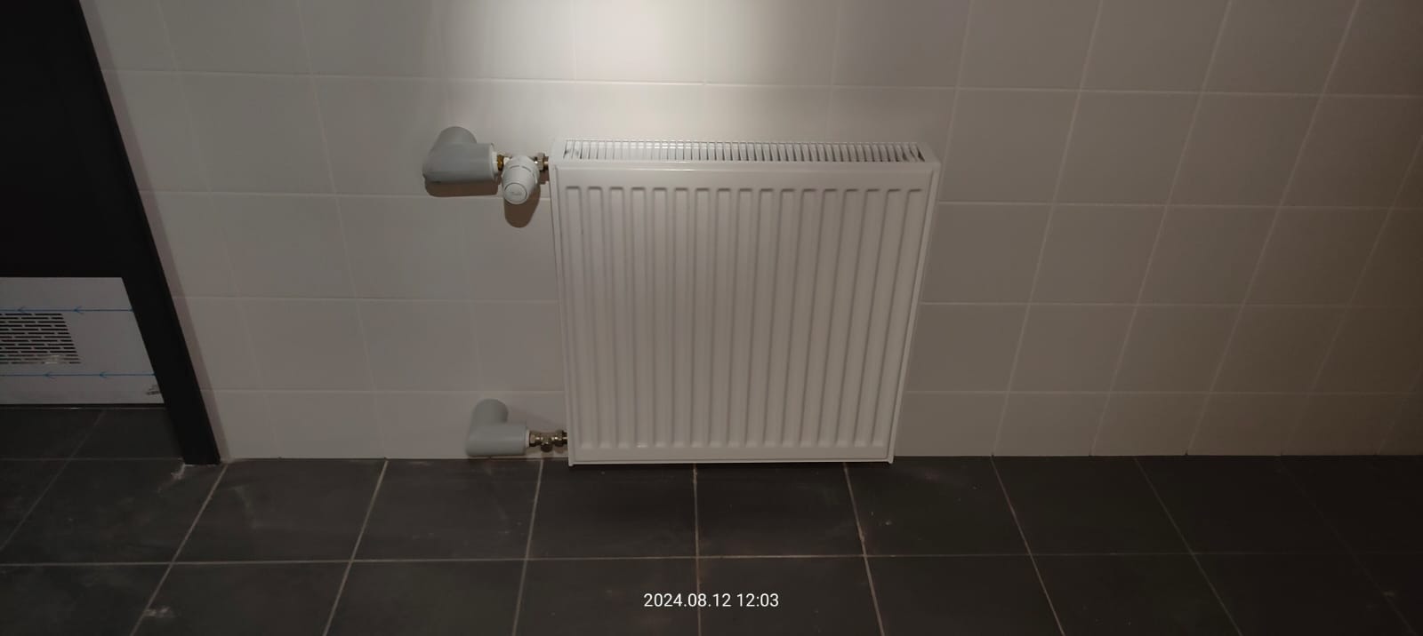 Biały grzejnik panelowy zamontowany na ścianie wyłożonej jasnymi płytkami, z widocznymi przyłączeniami i termostatem, na tle ciemnej podłogi z płytek.