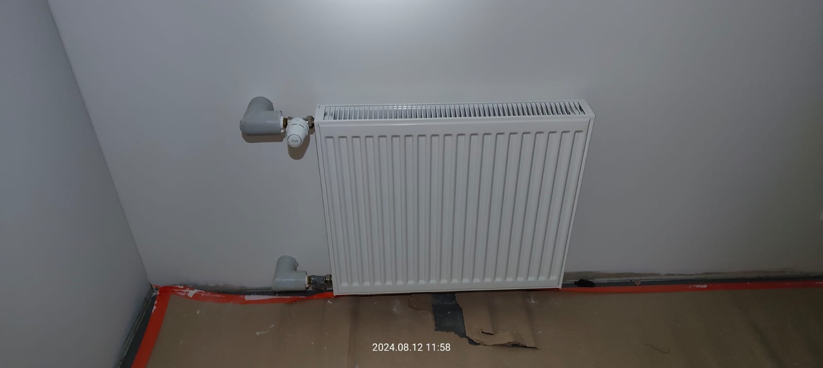 Biały grzejnik panelowy z termostatem zamontowany na ścianie, widoczne rury doprowadzające, podłoga zabezpieczona kartonem i taśmą malarską.