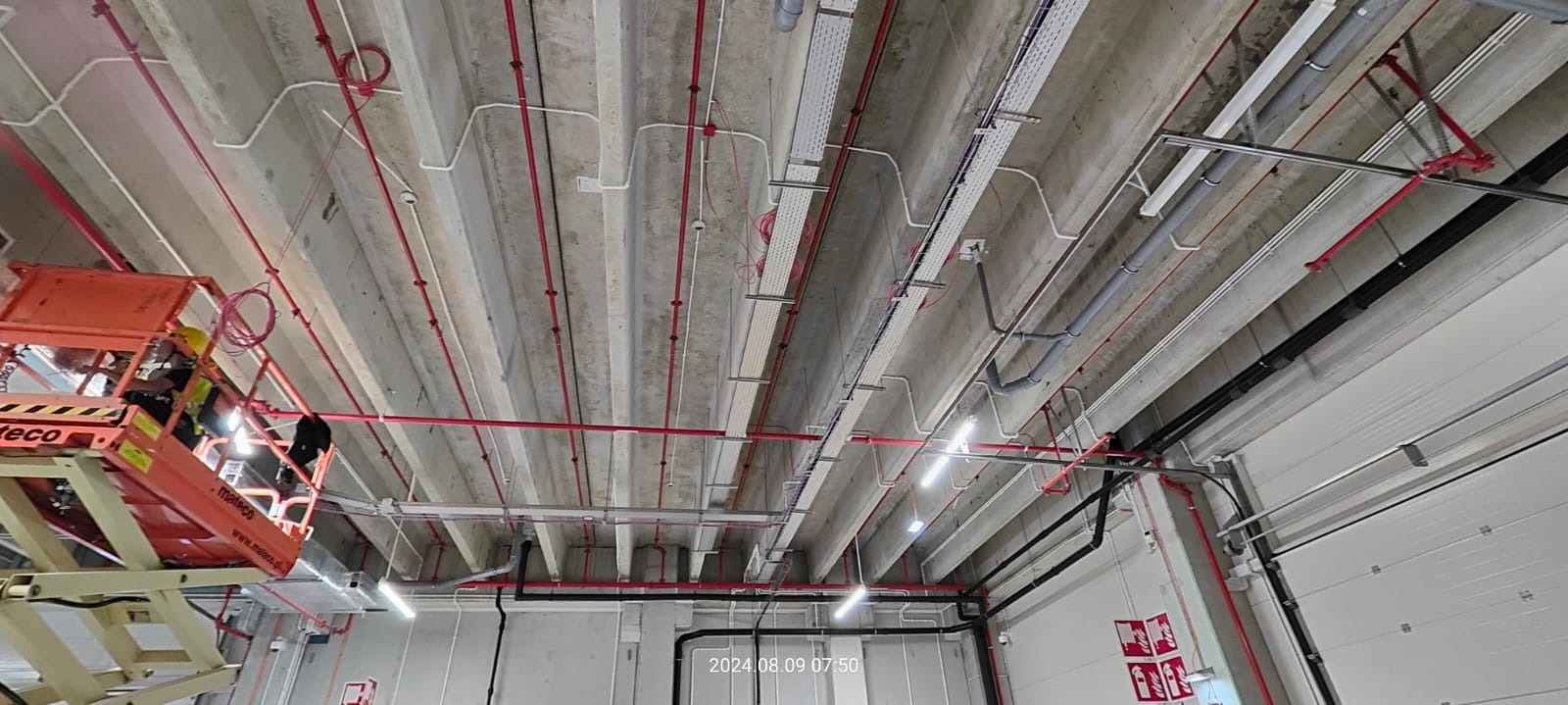 Montaż instalacji pod sufitem w hali przemysłowej, widoczna platforma podnośnikowa z pracownikami, liczne kable i rury w kolorach czerwonym, białym i czarnym, oraz metalowe korytka kablowe.