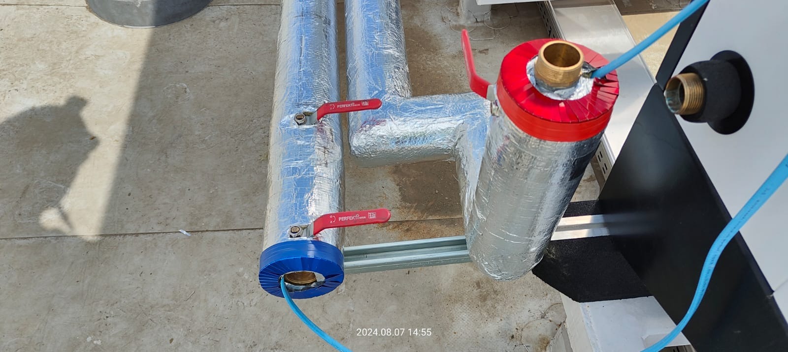 Ocieplone rury instalacji grzewczej z zaworami odcinającymi, zakończone niebieską i czerwoną osłoną termiczną, połączone niebieskim przewodem.