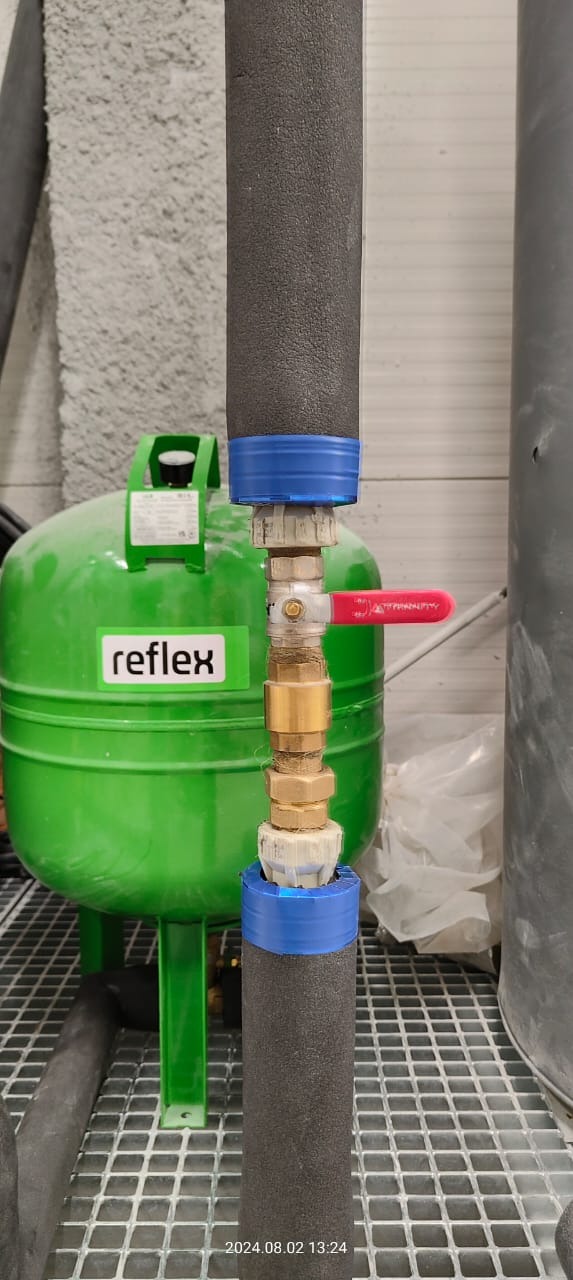 Zielony zbiornik wyrównawczy Reflex z podłączonymi rurami izolowanymi termicznie, połączonymi armaturą z czerwoną dźwignią zaworu, widoczne niebieskie pierścienie mocujące izolację.
