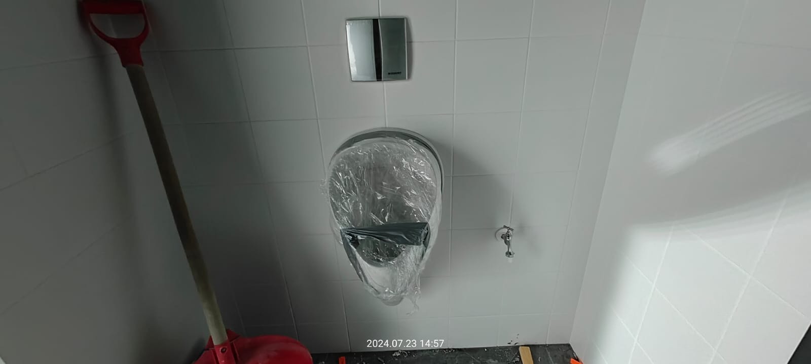 Nowy pisuar z osłoną z folii ochronnej, zamontowany na białej, kafelkowanej ścianie. Widoczny przycisk spłuczki. Po lewej oparta o ścianę szpadel z czerwonym uchwytem. Na podłodze resztki po remoncie.