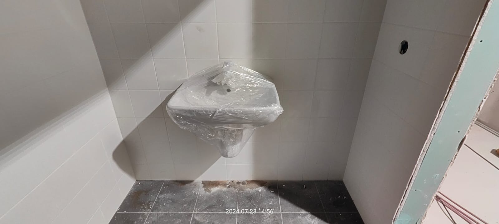 Umywalka na postumencie, zabezpieczona folią ochronną, w trakcie prac wykończeniowych łazienki. Widoczne białe płytki ścienne, szara podłoga i surowa ściana z płyty gipsowo-kartonowej.