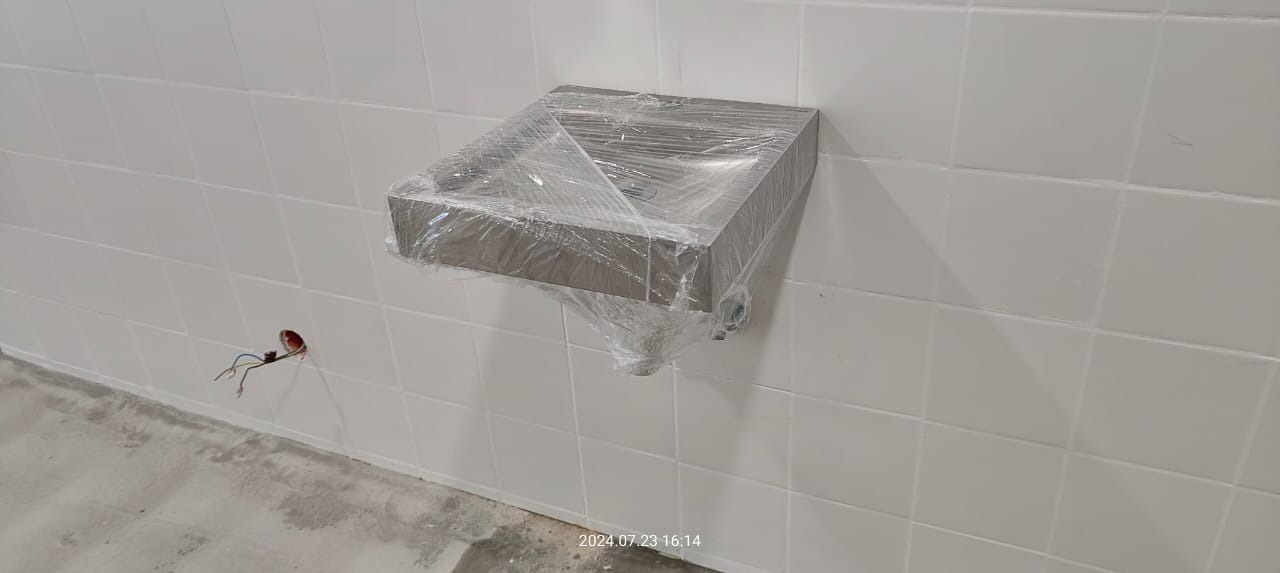 Nowa, prostokątna umywalka z szarego konglomeratu, zabezpieczona folią ochronną, zamontowana na białej ścianie z płytek ceramicznych. Widoczny wystający kabel elektryczny w puszce.