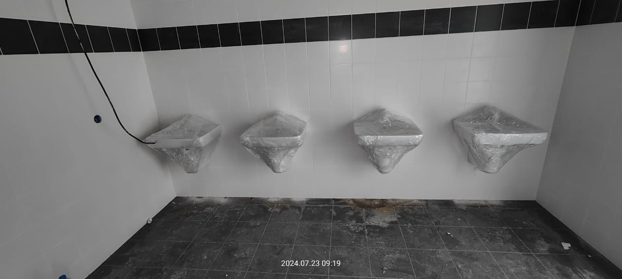 Cztery nowe, kwadratowe umywalki, osłonięte folią ochronną, zamontowane na ścianie wyłożonej białymi płytkami z czarnym pasem dekoracyjnym pod sufitem, podłoga wyłożona ciemnymi płytkami w trakcie...