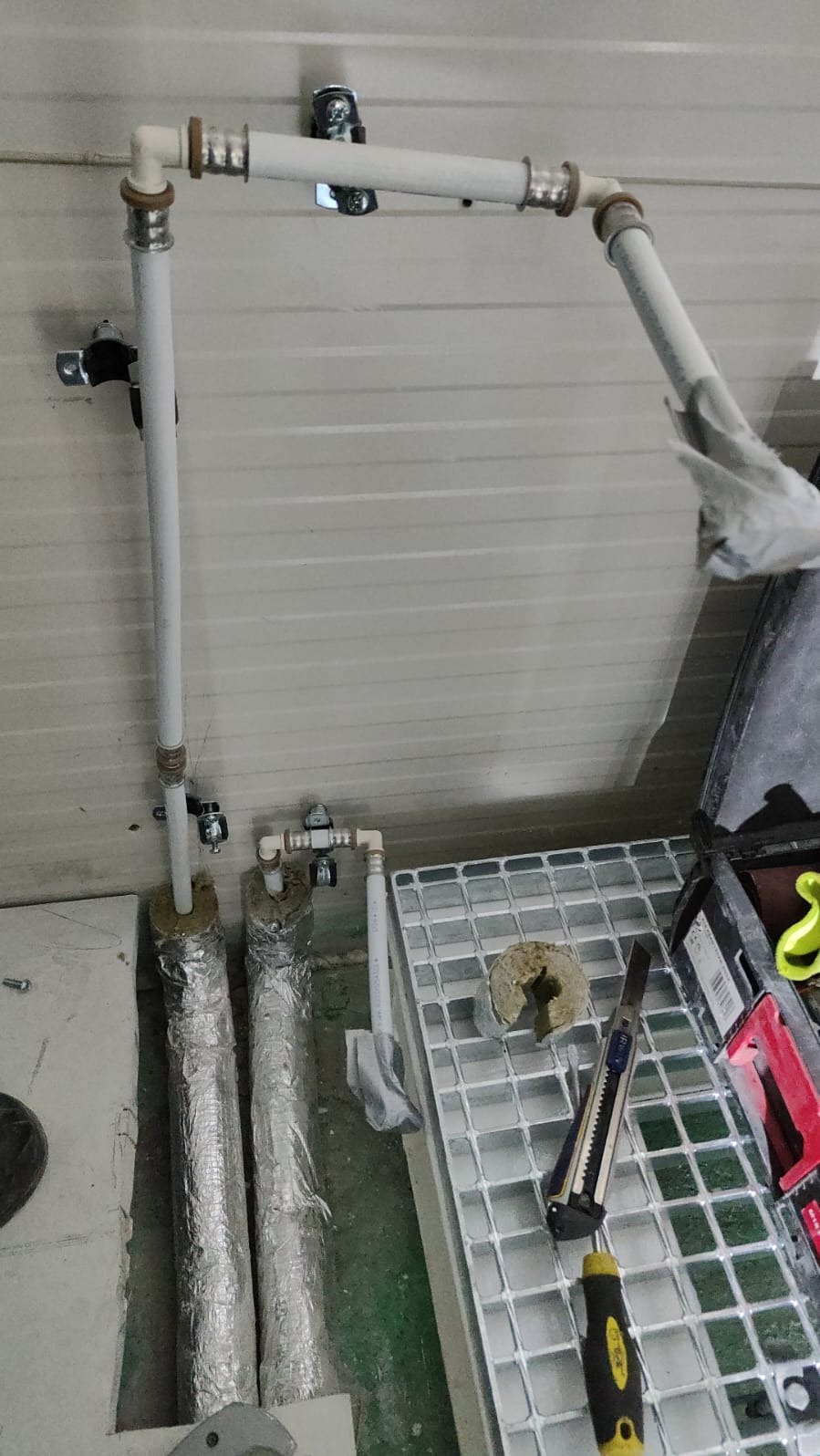 Instalacja rur grzewczych z izolacją termiczną, widoczne narzędzia: nóż monterski i śrubokręt, a także skrzynka narzędziowa w tle.