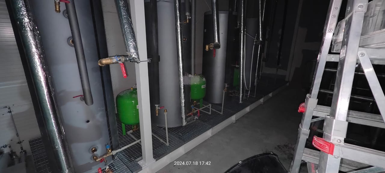 Rząd szarych i czarnych zbiorników ciśnieniowych oraz zielonych zbiorników wyrównawczych, połączonych rurami i zaworami, ustawionych na metalowej platformie pod ścianą. Z prawej strony widoczny...