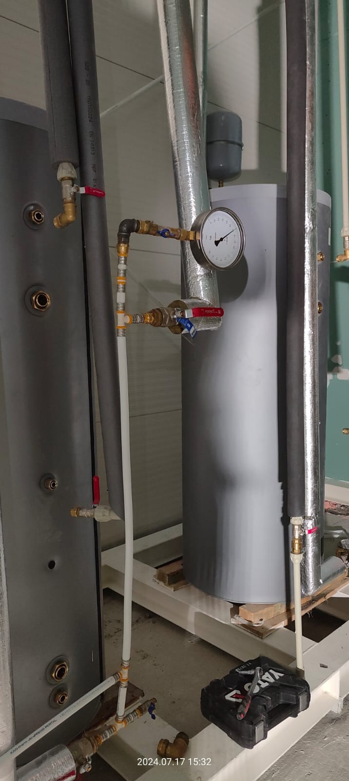 Szare zbiorniki akumulacyjne ciepła z rurami, zaworami i manometrem, instalacja na białej konstrukcji stalowej, skrzynka z narzędziami Yato na podłodze.