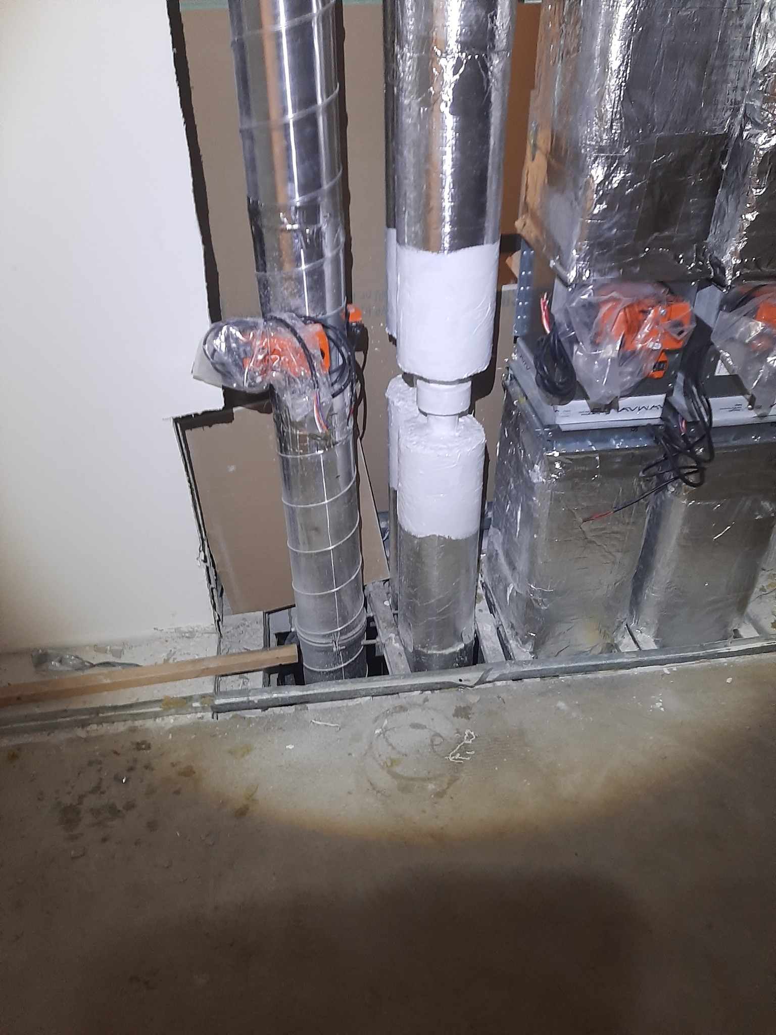 Fragment instalacji z rurami owiniętymi folią aluminiową oraz białym materiałem izolacyjnym, widoczne pomarańczowe elementy, podłoga w stanie surowym.