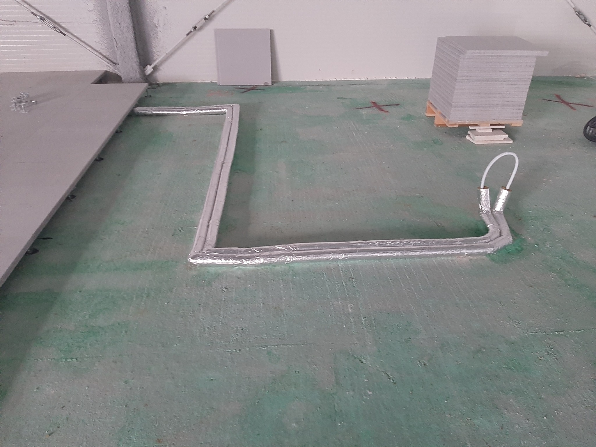 Izolowane rury ułożone na zielonej podłodze w kształcie litery U, z paletą płyt w tle i elementami konstrukcyjnymi na suficie.