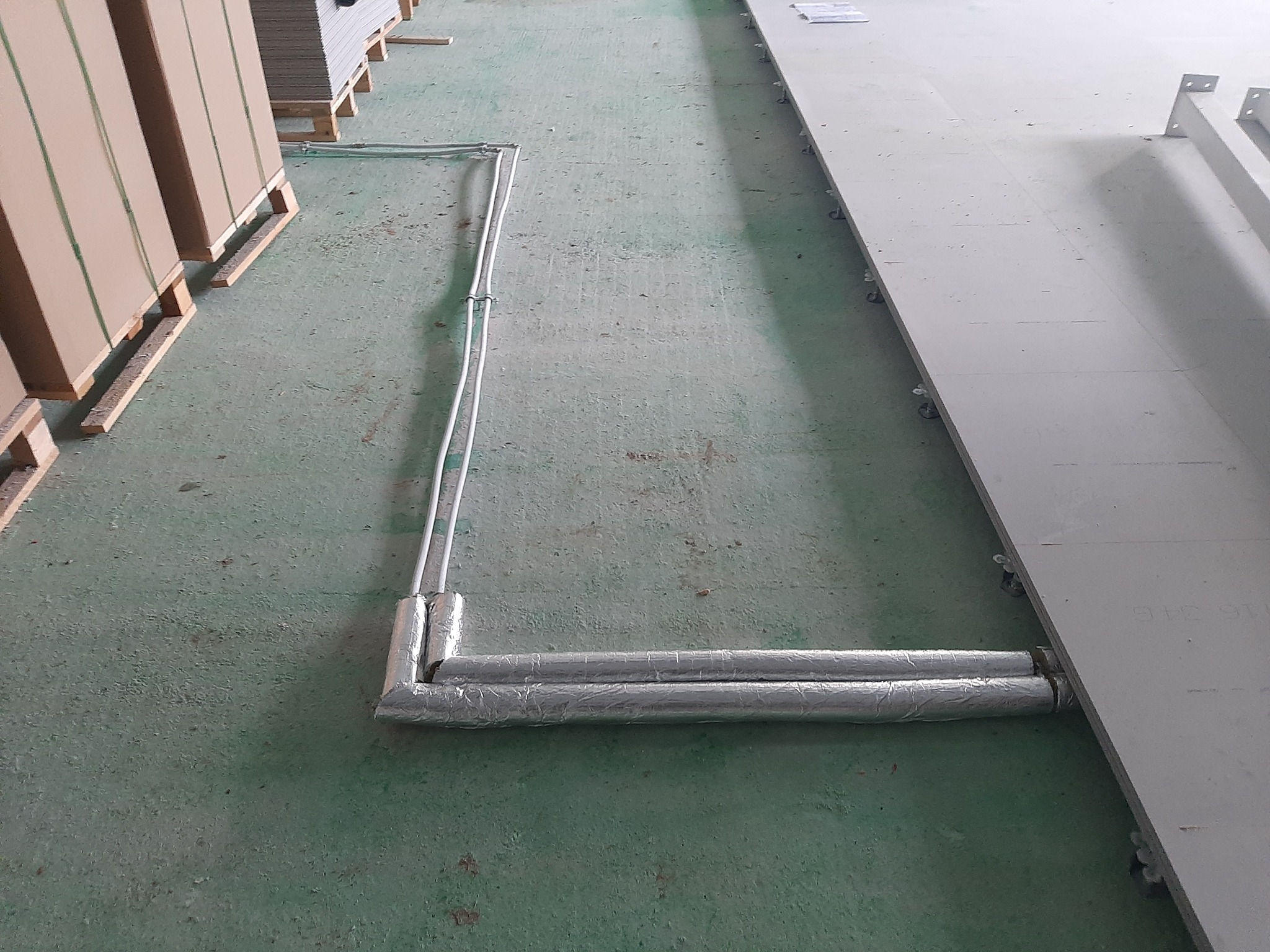 Dwie rury równoległe, owinięte srebrną folią izolacyjną, biegnące wzdłuż ściany w pomieszczeniu o zielonej podłodze. Rury połączone z pionowym przewodem. Palety z płytami w tle.