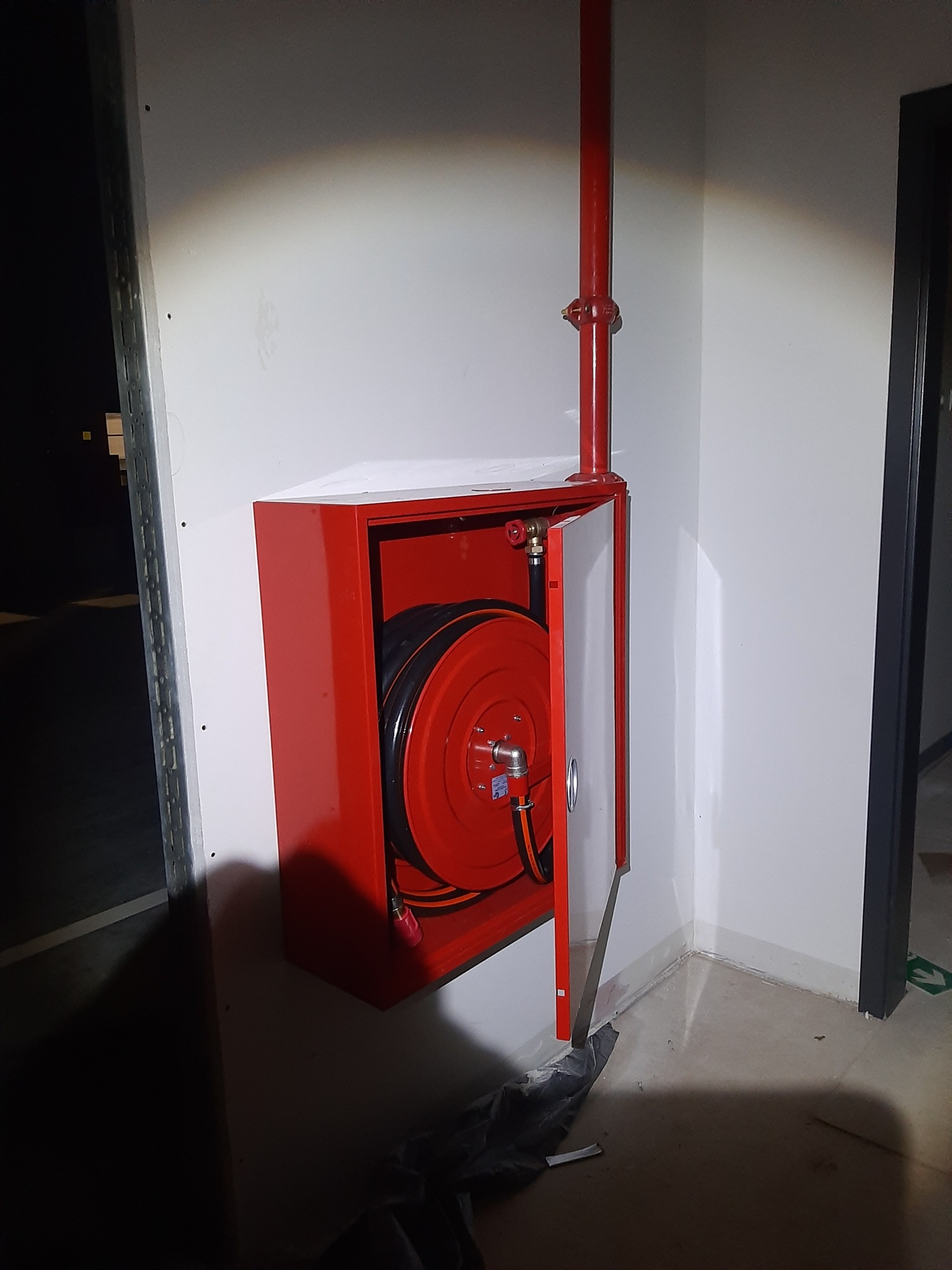 Czerwona szafka hydrantowa z rozłożonym wężem na ścianie wewnątrz budynku, z widoczną rurą doprowadzającą wodę.