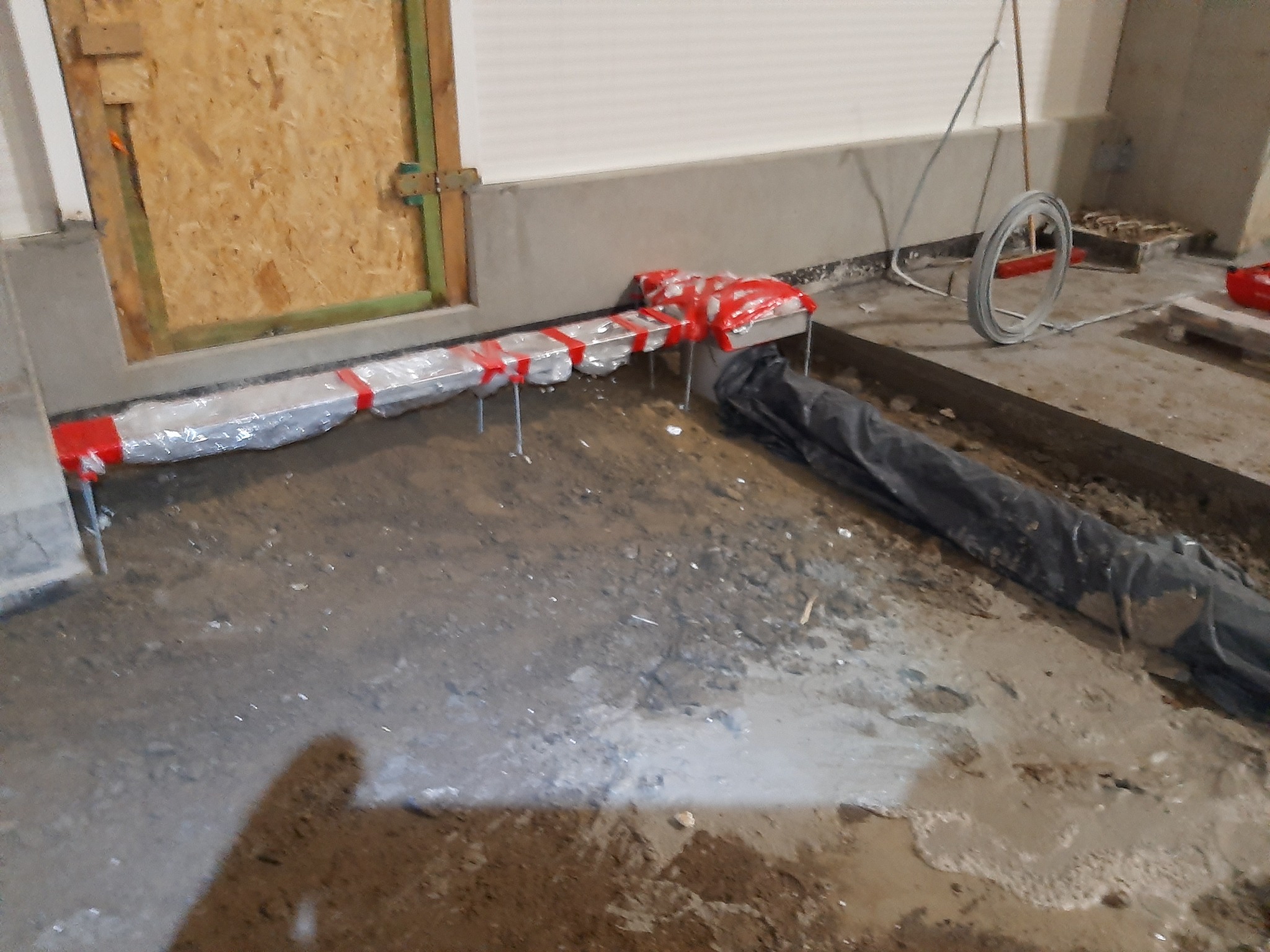 Przygotowanie podłoża pod instalację grzewczą: rury owinięte folią i zabezpieczone czerwoną taśmą, wystające metalowe wsporniki, czarna folia ochronna na podłodze w trakcie prac.
