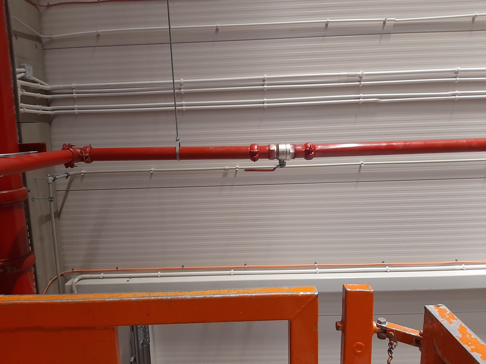 Czerwona rura instalacji przeciwpożarowej z zaworem odcinającym, przymocowana do sufitu za pomocą metalowych obejm. Nad nią widoczne białe rury instalacyjne. W dolnej części kadru pomarańczowa...