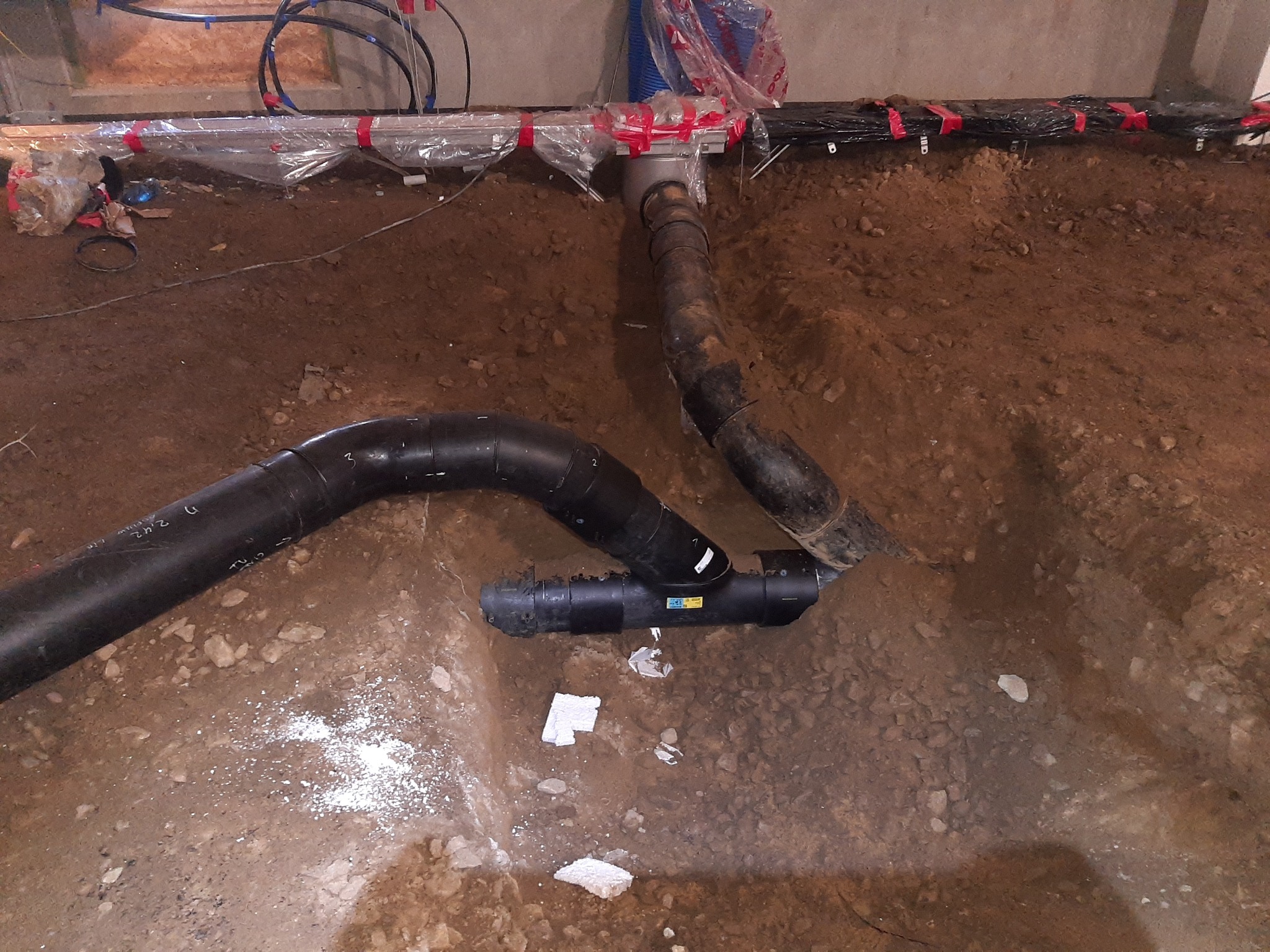 System rur kanalizacyjnych z tworzywa sztucznego, częściowo zakopany w ziemi, widoczne połączenia i mocowania, elementy instalacji zabezpieczone folią.