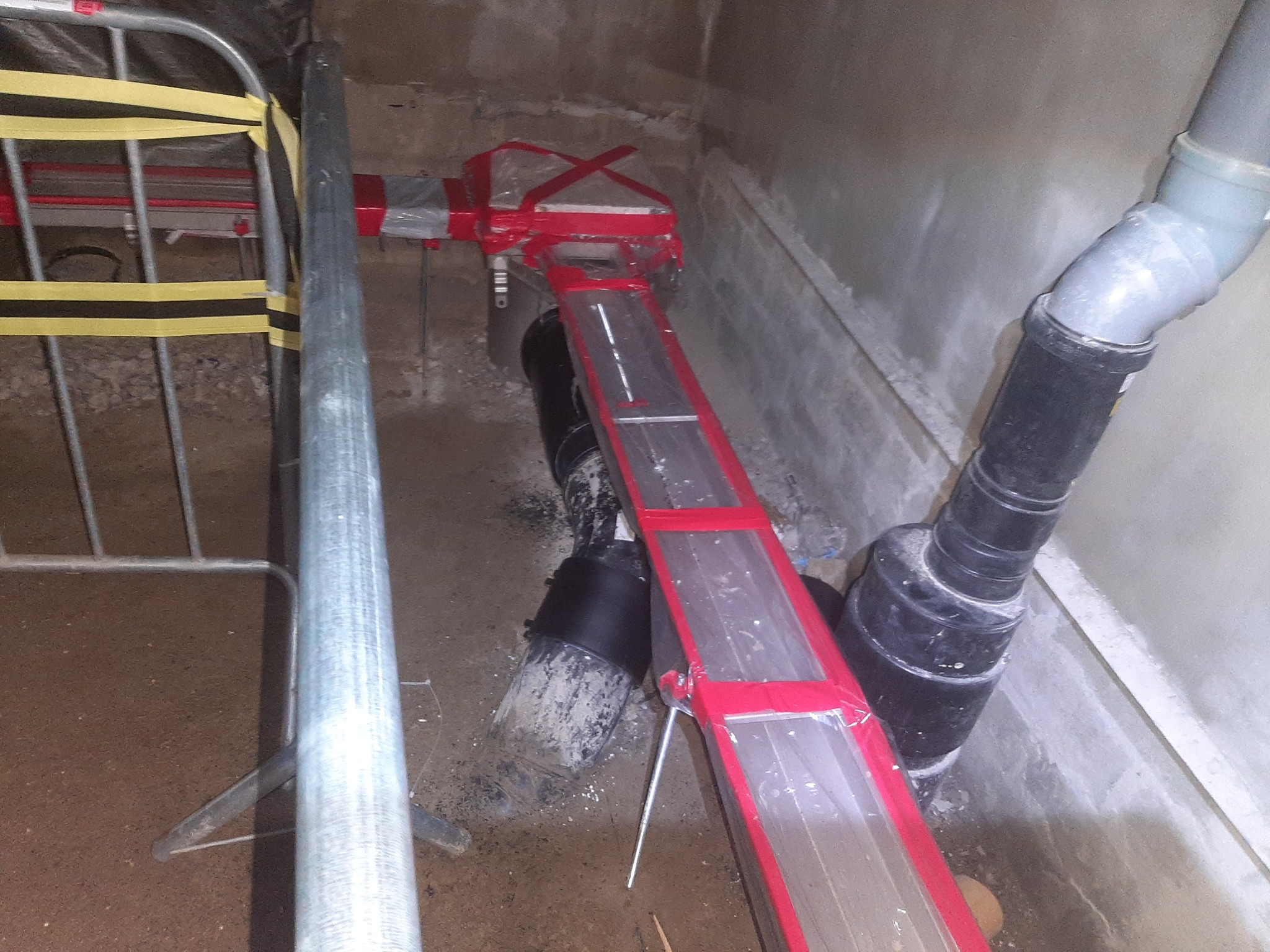 Fragment instalacji kanalizacyjnej z rurami PCV, zabezpieczonej czerwoną konstrukcją wsporczą z folią, w trakcie prac w surowym stanie.