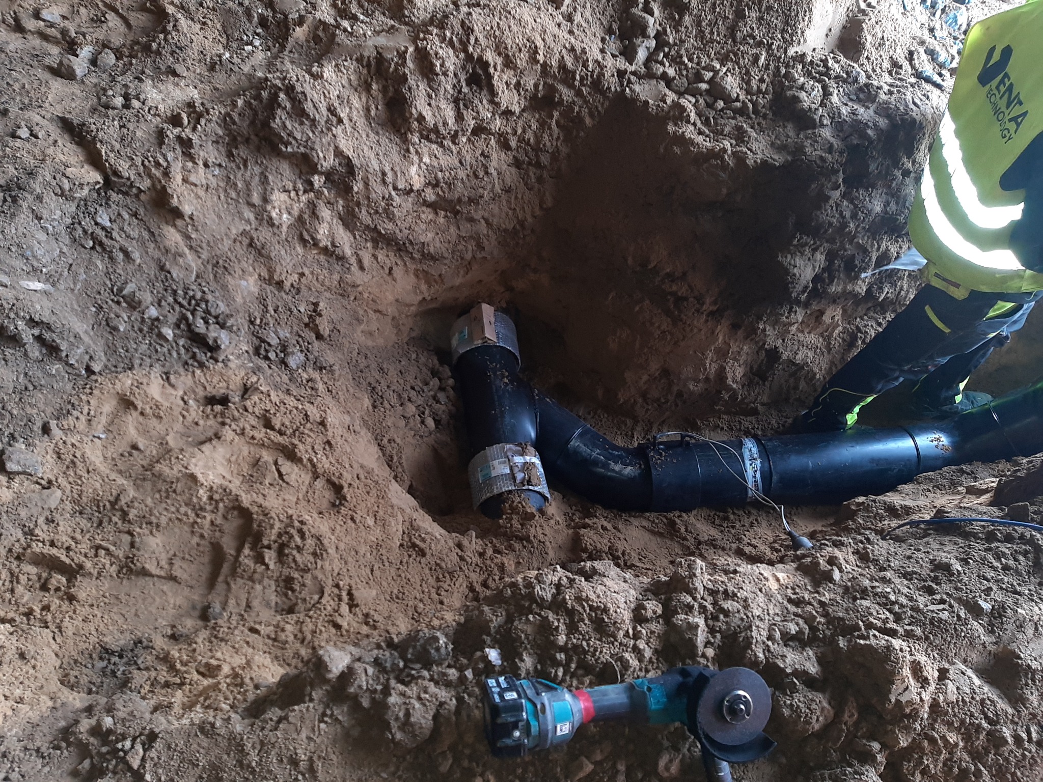 Czarny rurociąg z łącznikami w wykopie ziemnym, obok leży szlifierka kątowa, widoczny fragment pracownika w odblaskowej kamizelce.