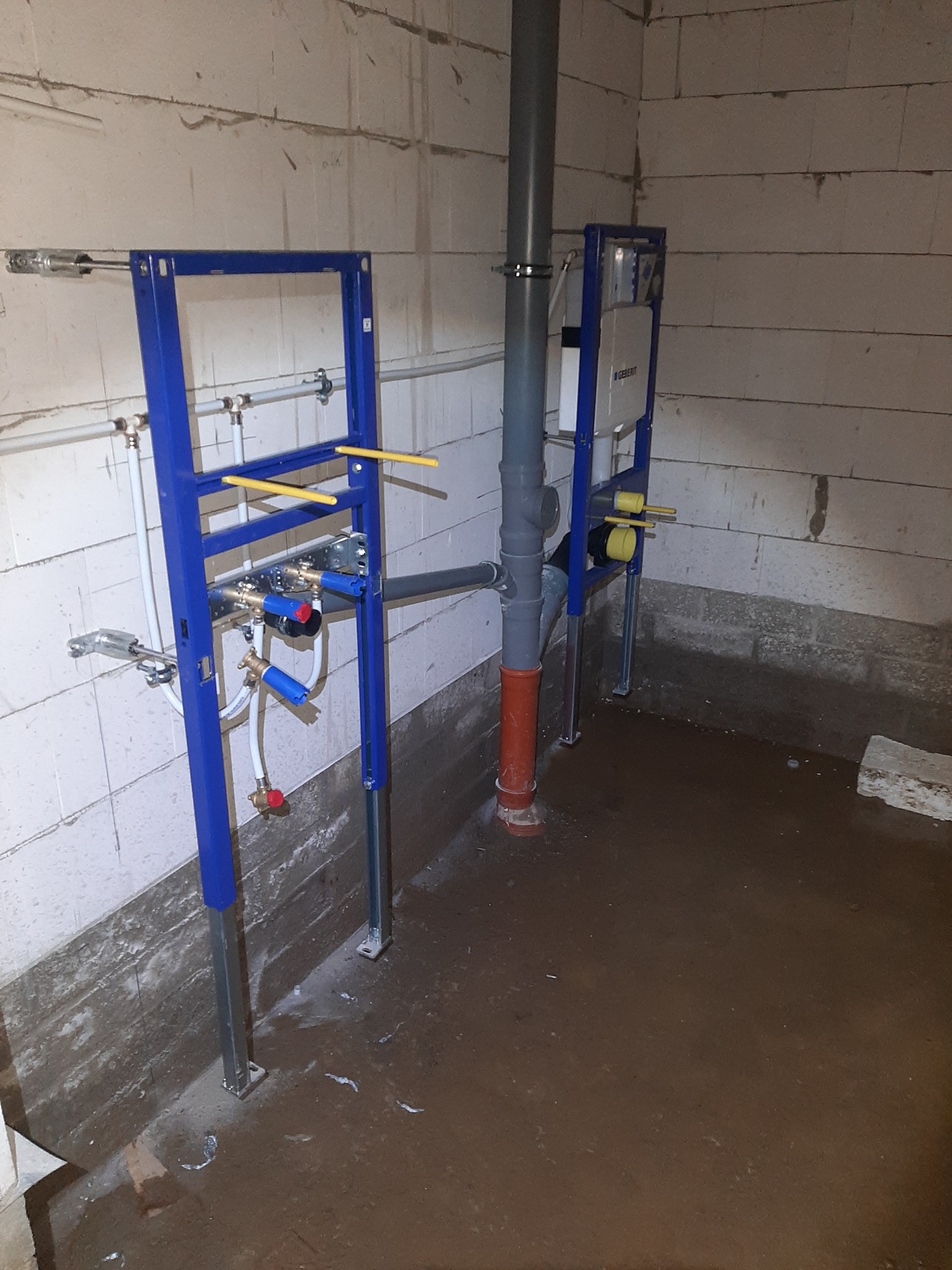 Stelaże podtynkowe WC z instalacją hydrauliczną w surowym stanie łazienki, widoczne rury odpływowe i doprowadzające wodę, ściany z białego bloczka.