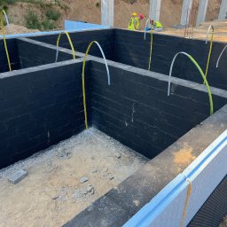 B_P ELEKTROINSTALACJE BARTOSZ PUCZKA - Fundamenty budynku z izolacją termiczną, czarną hydroizolacją i wyprowadzonymi przewodami elektrycznymi, w tle pracownicy w kamizelkach odblaskowych.