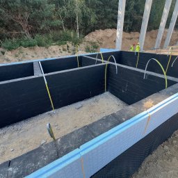 B_P ELEKTROINSTALACJE BARTOSZ PUCZKA - Fundamenty budynku z izolacją termiczną, czarną hydroizolacją i wystającymi przewodami elektrycznymi, widoczni pracownicy w oddali.