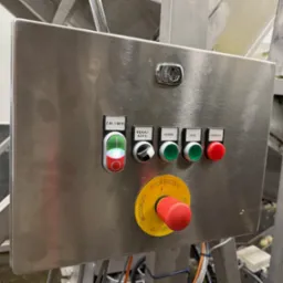 Panel sterowania ze stali nierdzewnej z przyciskami (włącz/wyłącz, ręka/auto, góra, dół, awaria) oraz wyłącznikiem bezpieczeństwa z żółtą obwódką, zamontowany na maszynie przemysłowej.