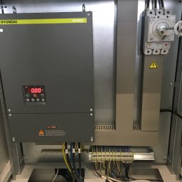 B_P ELEKTROINSTALACJE BARTOSZ PUCZKA - Wnętrze szafy sterowniczej z zamontowanym falownikiem Hyundai N700E, wyłącznikiem Eaton NZM2 oraz listwą zaciskową z podłączonymi przewodami. Widoczne oznaczenia ostrzegawcze o ryzyku porażenia...