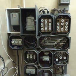 B_P ELEKTROINSTALACJE BARTOSZ PUCZKA - Rozdzielnia elektryczna z otwartymi obudowami, widoczne bezpieczniki topikowe, liczniki energii elektrycznej oraz okablowanie w starym budynku.