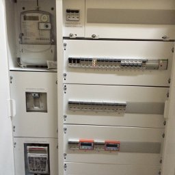 B_P ELEKTROINSTALACJE BARTOSZ PUCZKA - Otwarta szafa elektryczna z licznikiem głównym, wyłącznikiem głównym, zabezpieczeniem przedlicznikowym i szeregiem wyłączników nadprądowych oraz modułów pomiarowych, widoczne oznaczenia numeryczne...