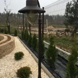 Rustykalna lampa ogrodowa z kloszem w kształcie dzwonu oświetla ścieżkę z białego żwiru, obok posadzone tuje, w tle ażurowe ogrodzenie.