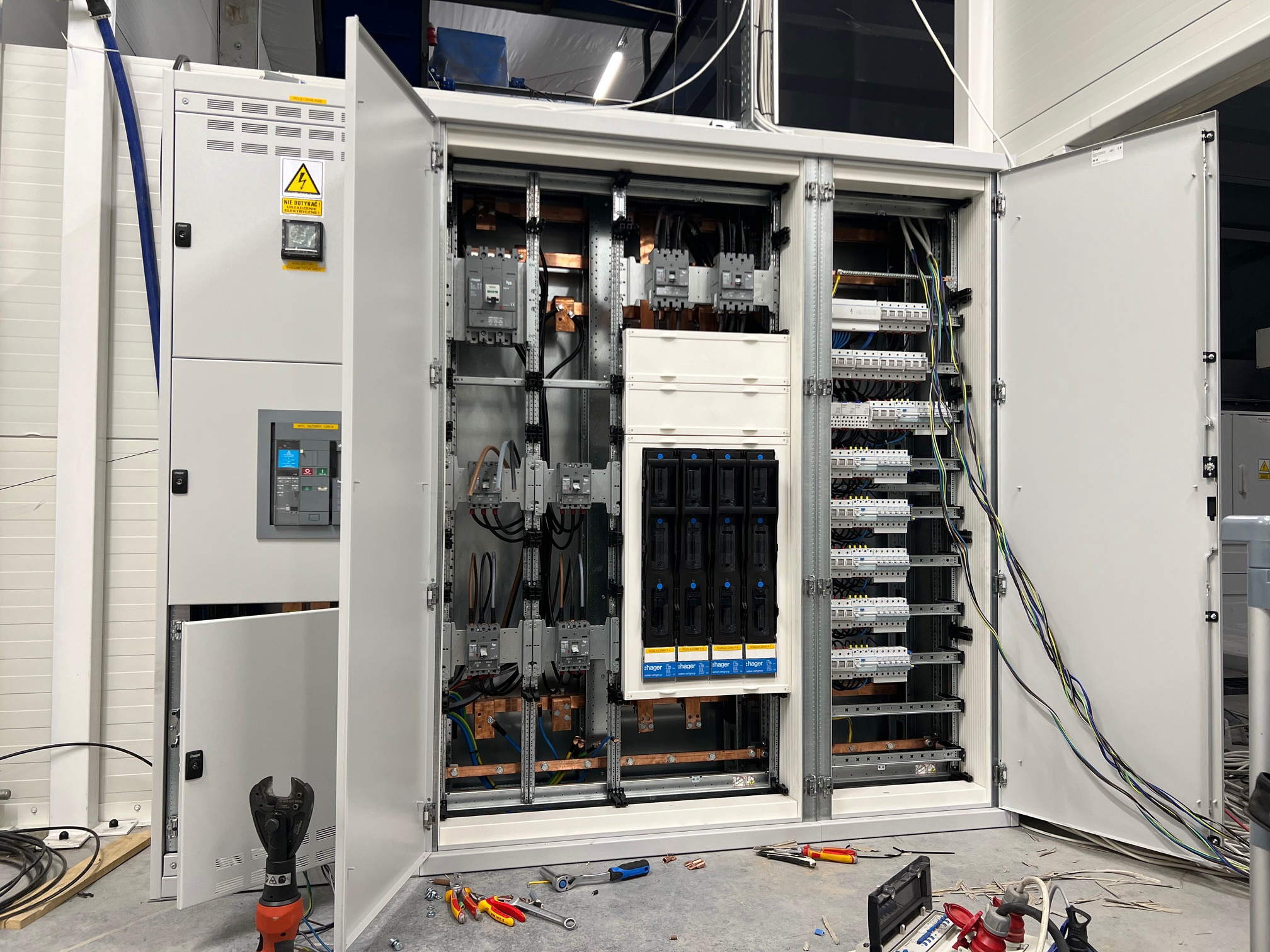 Otwarta szafa elektryczna z widocznymi elementami instalacji, takimi jak wyłączniki, bezpieczniki, kable i szyny miedziane. Na podłodze rozłożone narzędzia: kombinerki, klucze płaskie, obcinak...