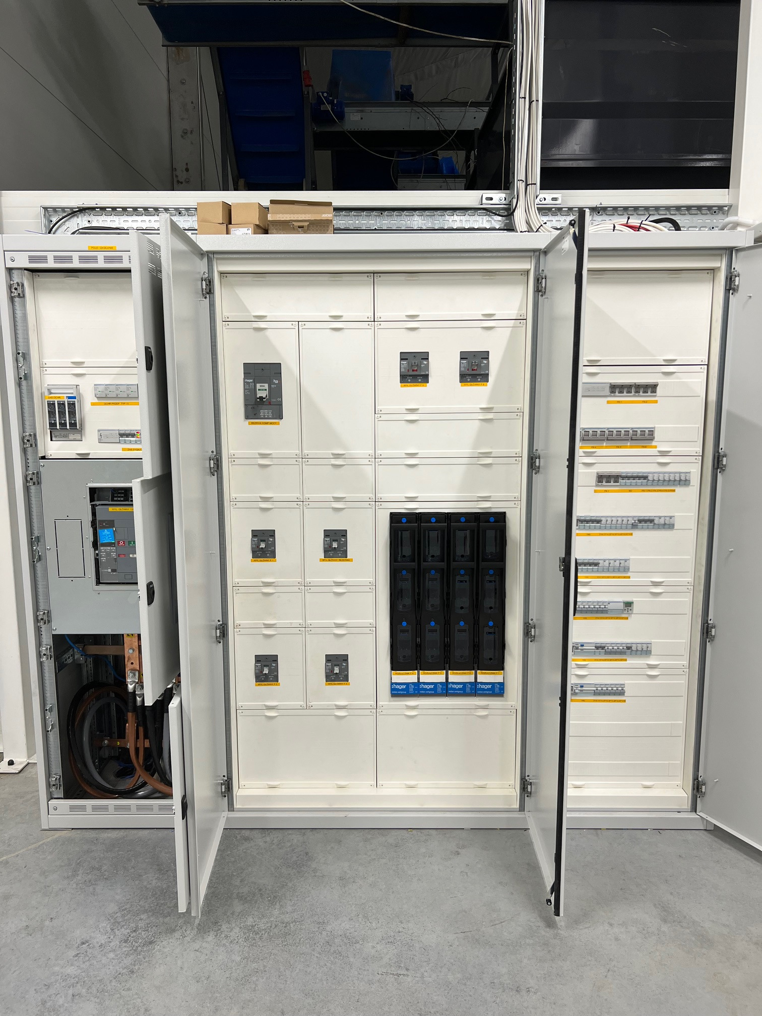 Otwarta, biała szafa rozdzielcza z widocznymi bezpiecznikami, przełącznikami i okablowaniem, umieszczona w przemysłowym wnętrzu z betonową podłogą.