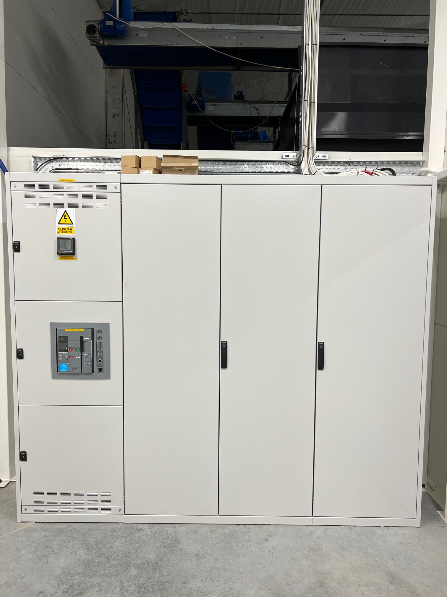 Rozdzielnica elektryczna w obudowie przemysłowej z widocznym wyłącznikiem głównym i miernikiem parametrów sieci, umieszczona w hali produkcyjnej.